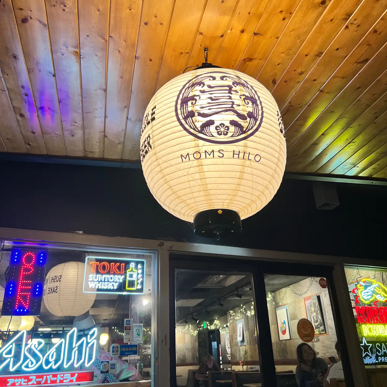 Illuminated white paper lantern with 'Moms Hilo' text hanging inside a restaurant, with neon signs and a wooden ceiling visible.