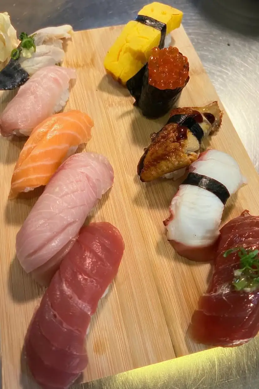 Assorted nigiri sushi on a wooden board including tuna, salmon, egg, octopus, eel with sauce, salmon roe, and white fish topped with garnishes.
