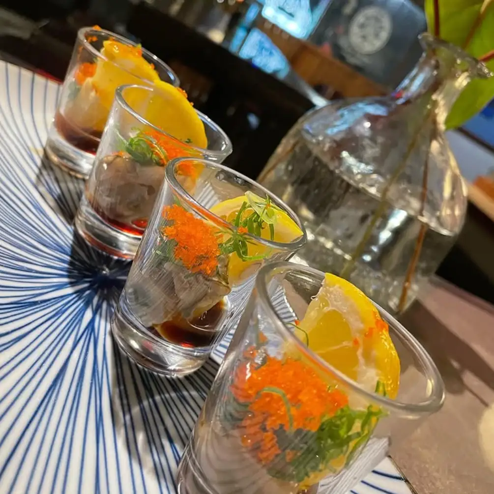 Four small glass cups arranged diagonally on a plate, each containing an oyster topped with orange fish roe, green garnish, and a lemon wedge.