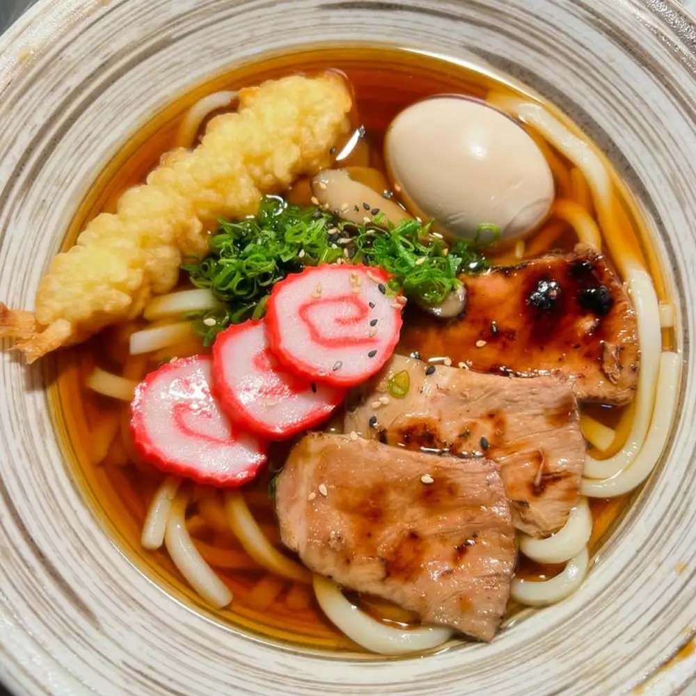 Bowl of udon soup with thick noodles, tempura shrimp, boiled egg, grilled meat slices, narutomaki fish cake, green onions, and sesame seeds in broth.